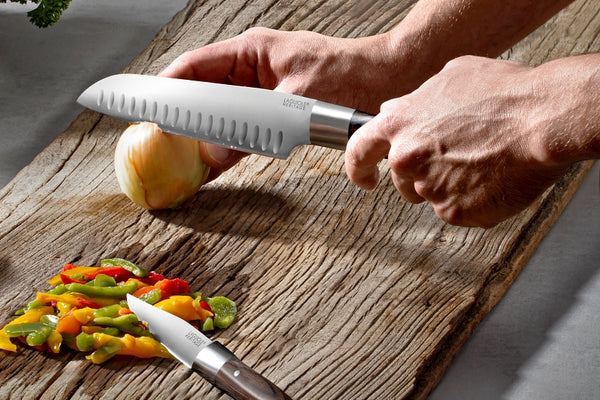 A person uses a Laguiole heritage pakkawood handle santoku knife to cut an onion. the background is composed of an old-looking piece of wood. 
