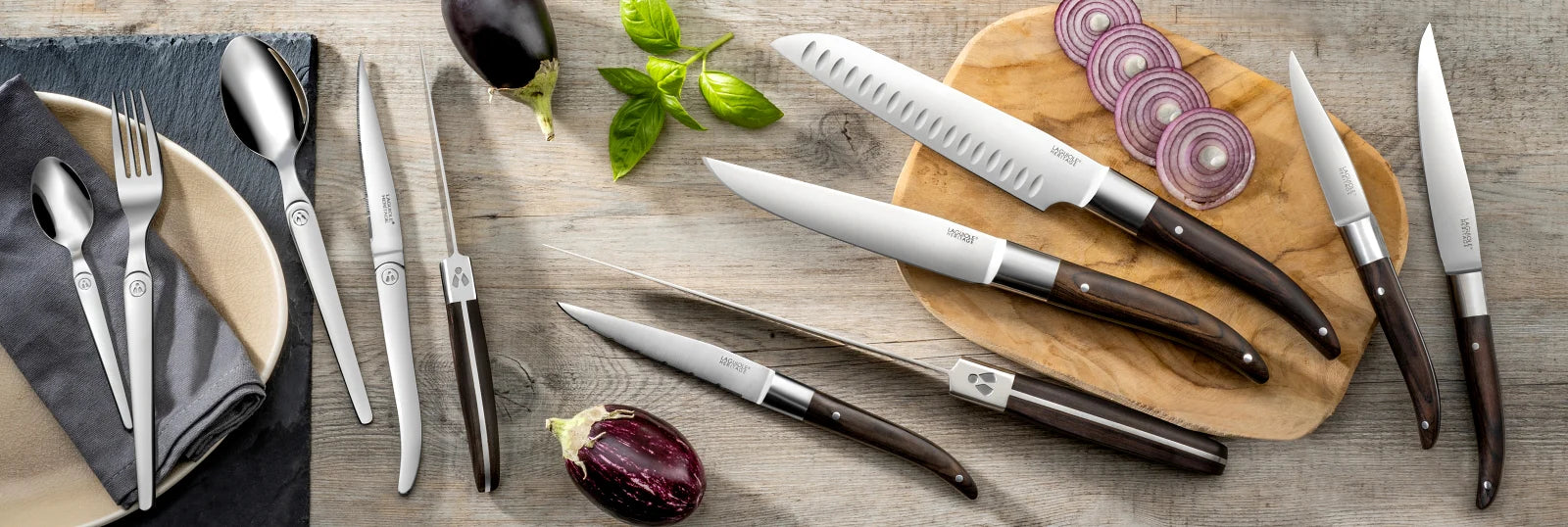 Several Laguiole heritage kitchen knives with wooden handles and flatware. The image background is made of light-colored wood.
